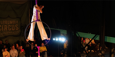A female teenage is standing on an Aerial Hoop high in the air looking down with an audience looking up at her