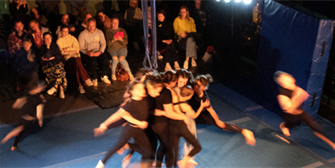 A group of youth acrobatics peform a merry go round with their bodies, another group circles them running and blurred in the image