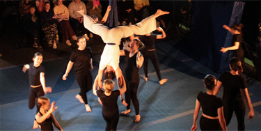 An aerialist dressed in white hangs upside down, lifting up a young girl, a group of acrobatic performers dressed in black circle the pair.