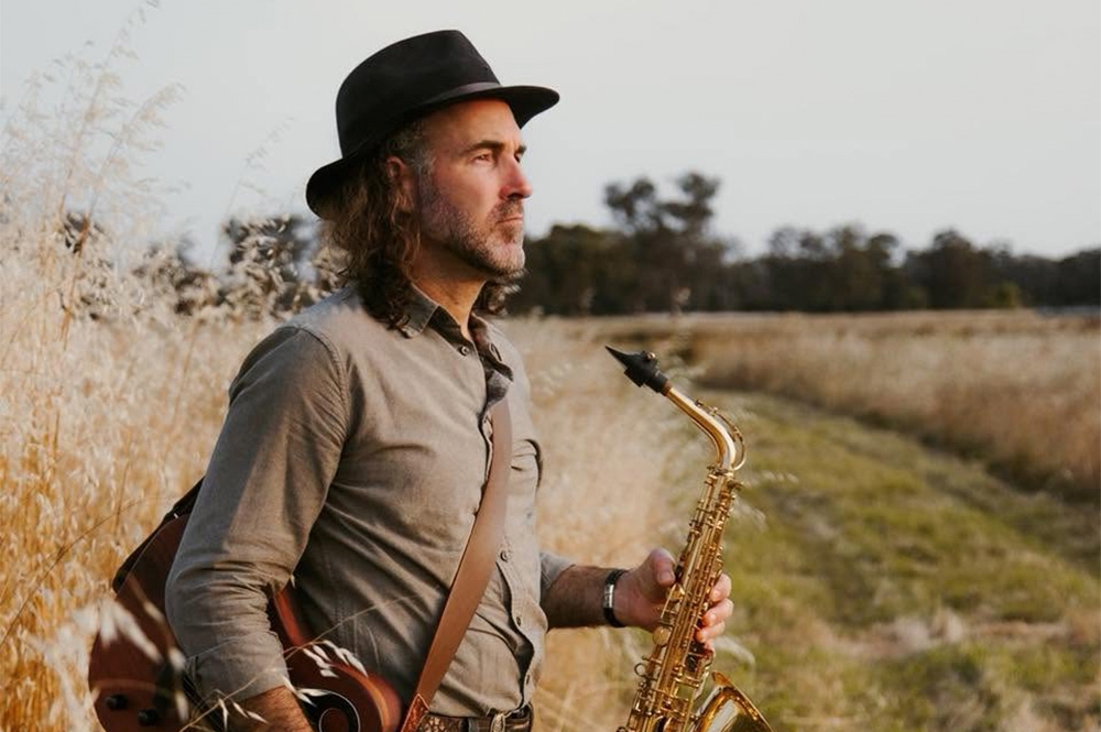 Dave Graffin standing outdoors in a rural field, wearing a black hat and holding a saxophone with a guitar visible behind him.